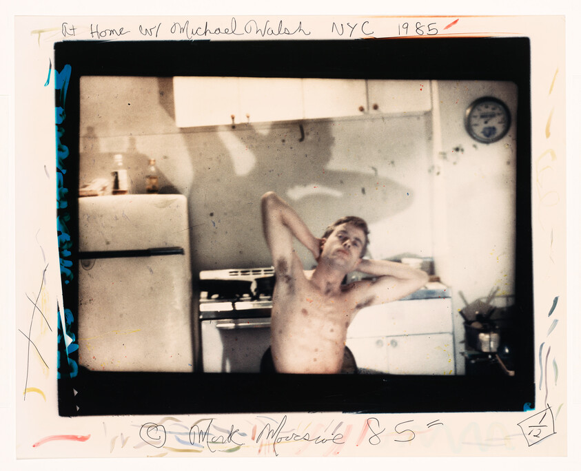 A shirtless man leans back with hands behind his head in a small kitchen.