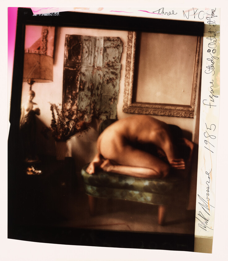 A nude person curled fetal on a small upholstered bench in a dimly lit room.