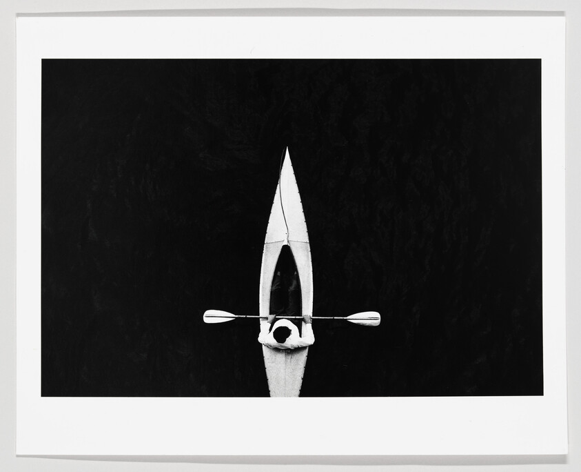 A person paddling a single white kayak across dark, still water, seen from above.