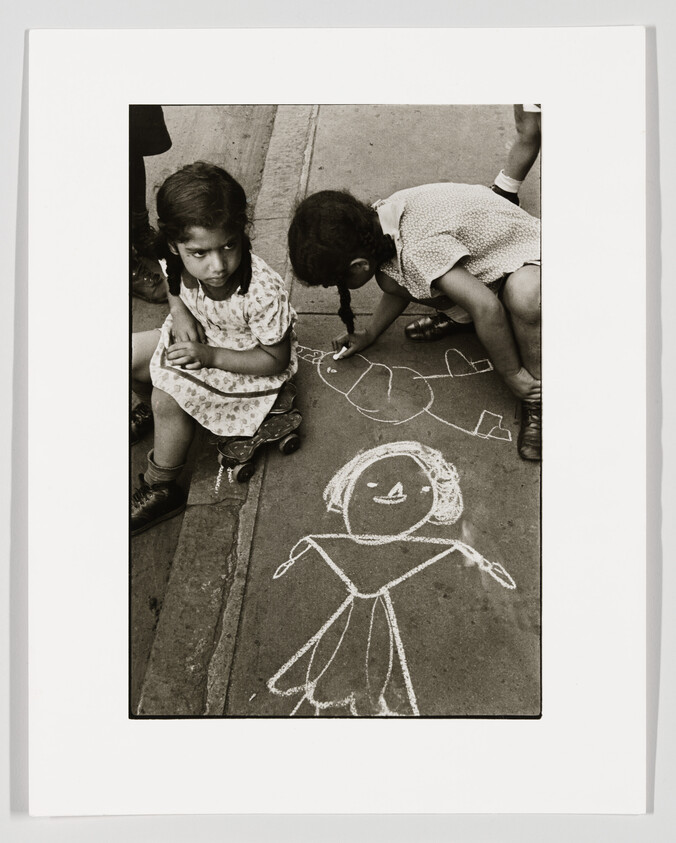 Two young girls draw chalk people on the sidewalk as one sits on a scooter.