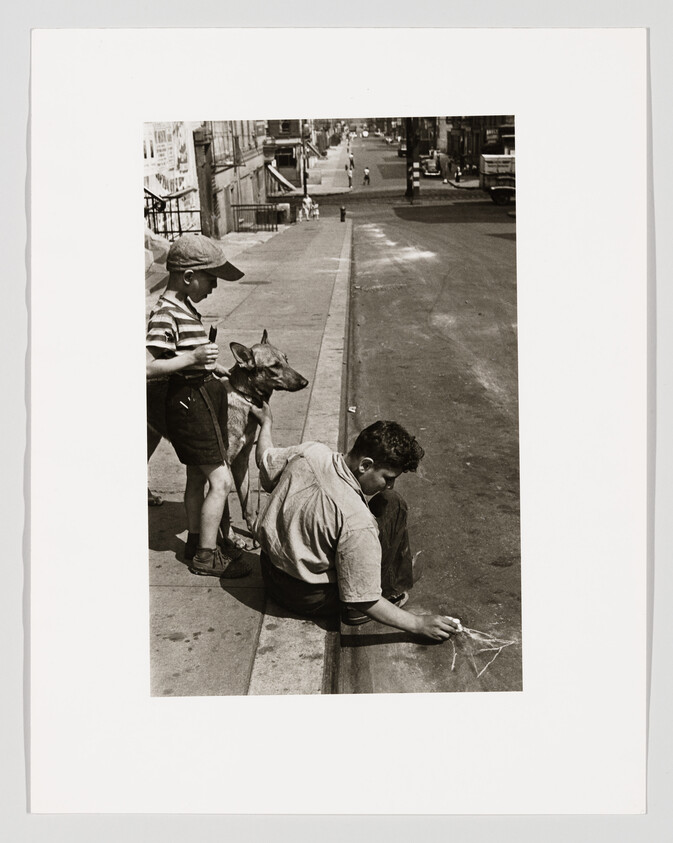 Two boys on a city curb, one sitting and drawing in the street while the other watches with a dog.