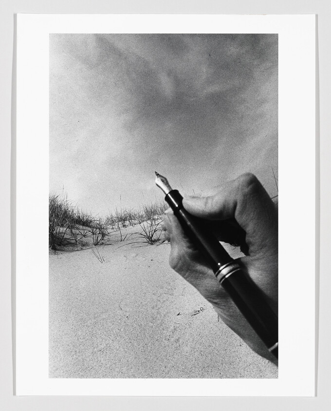 A hand holds a fountain pen pointing toward sandy dunes with sparse beach grass.