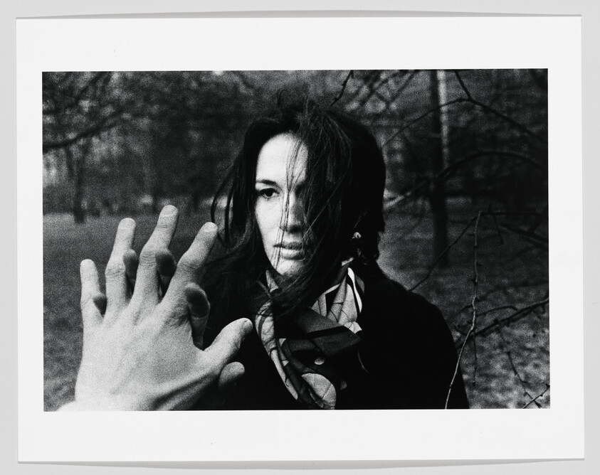 A hand reaches toward a woman with wind-blown hair standing in a bare park.