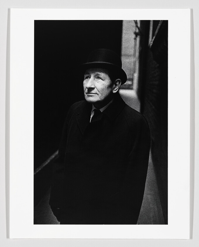 An elderly man in a bowler hat and dark coat looks upward in a narrow alley.