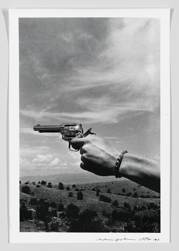 A hand wearing a bracelet holds a revolver pointed toward the sky over a dry landscape.