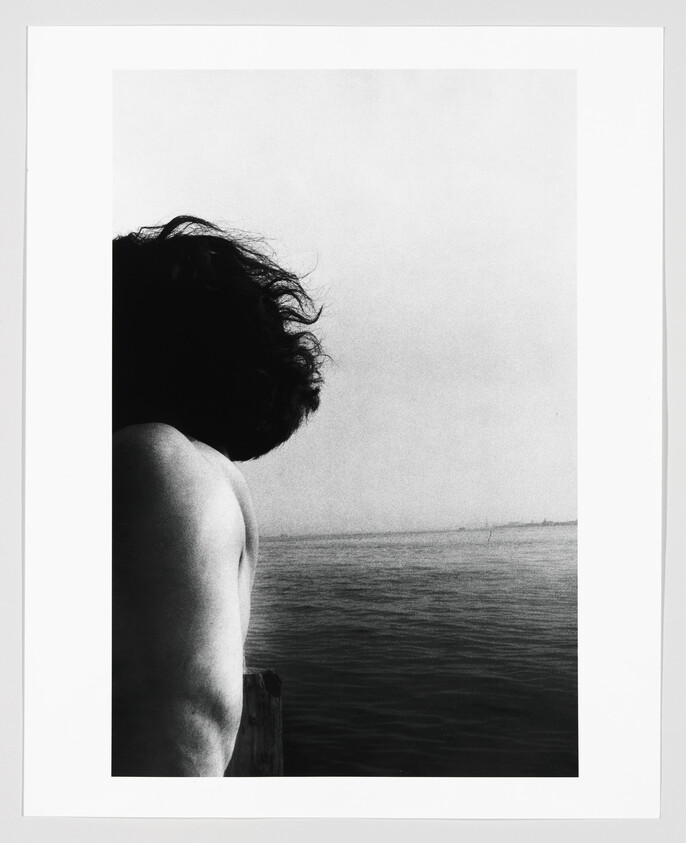 A shirtless person with tousled hair leans over a railing looking out across calm open water.