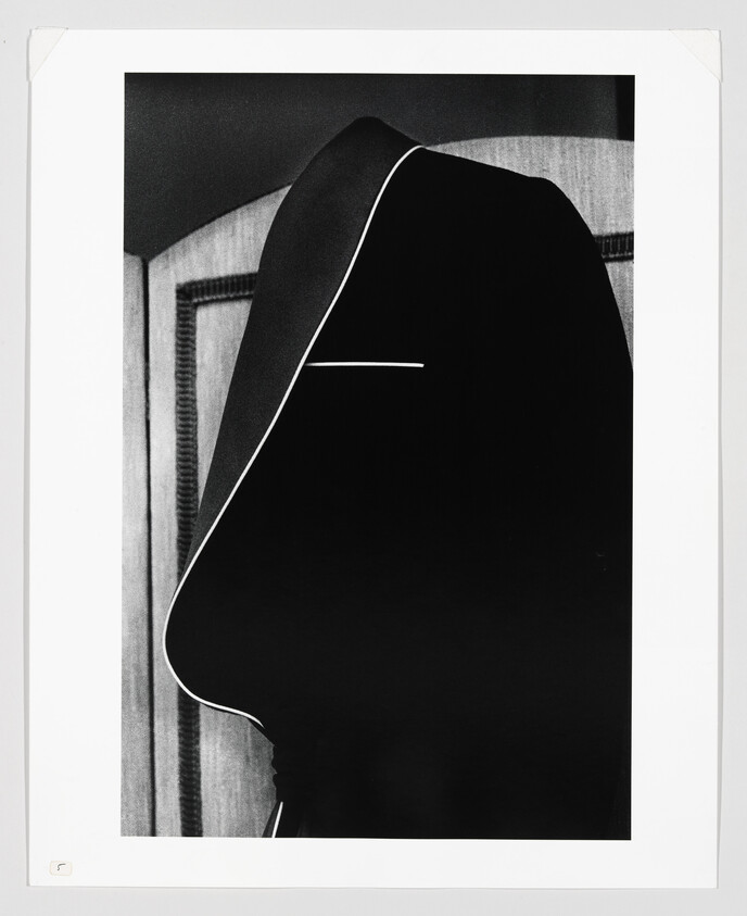 A person wearing a dark hooded cloak with white trim seen in profile against a paneled background.