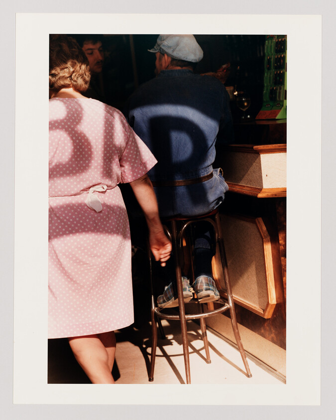 Woman in pink polka-dot dress walks past a man sitting at a bar under a large shadow.