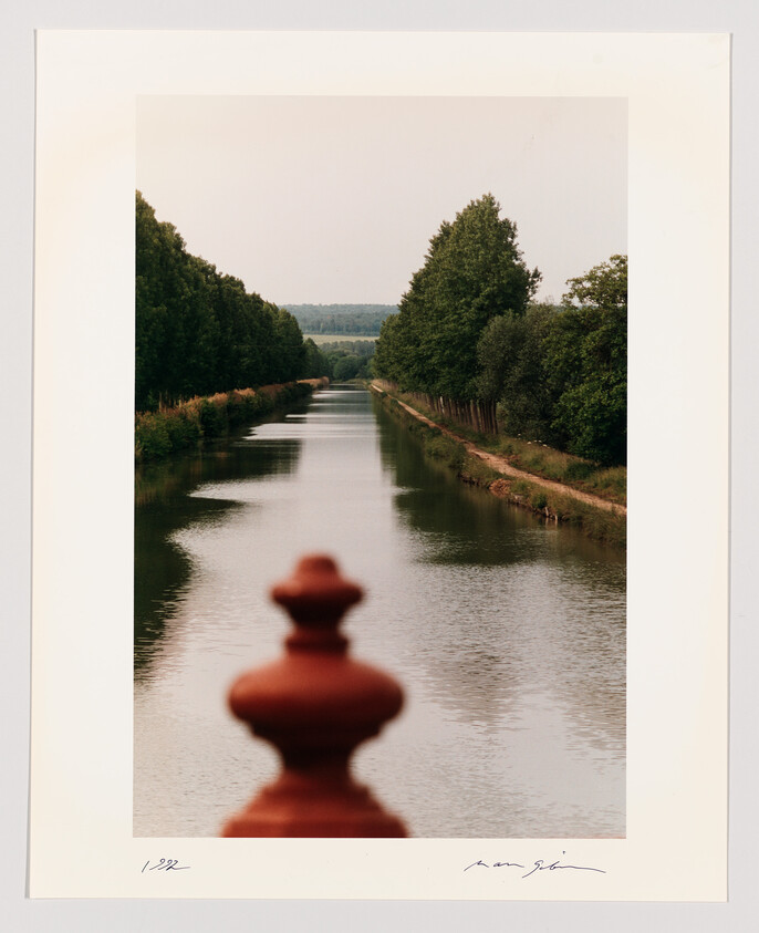 A quiet tree-lined canal stretches into the distance with a blurred red baluster in the foreground.