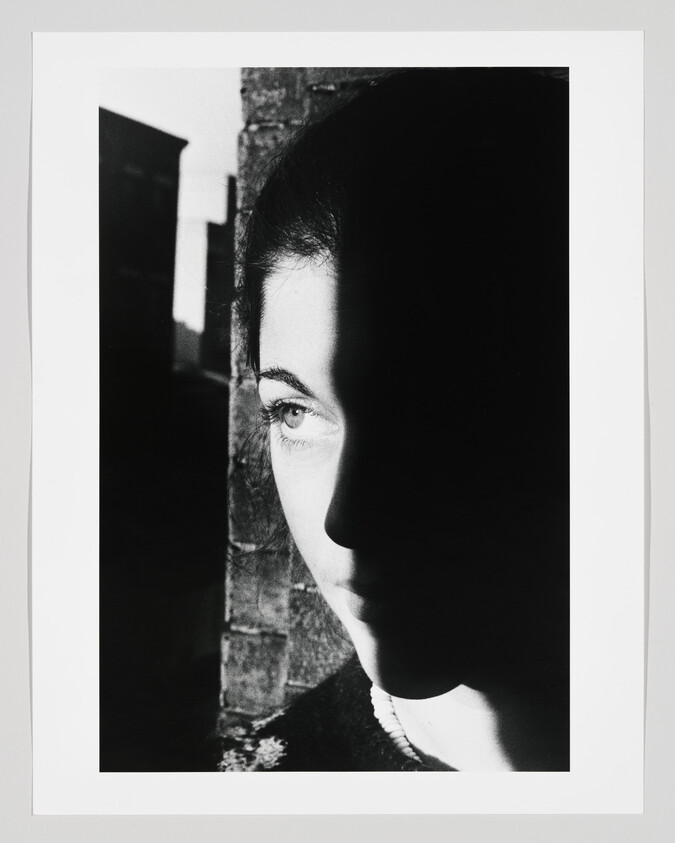 A woman's face half covered by shadow against a brick wall, one eye illuminated.