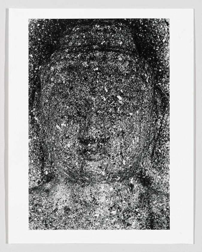Weathered stone statue face with worn features and speckled surface framed on white background.