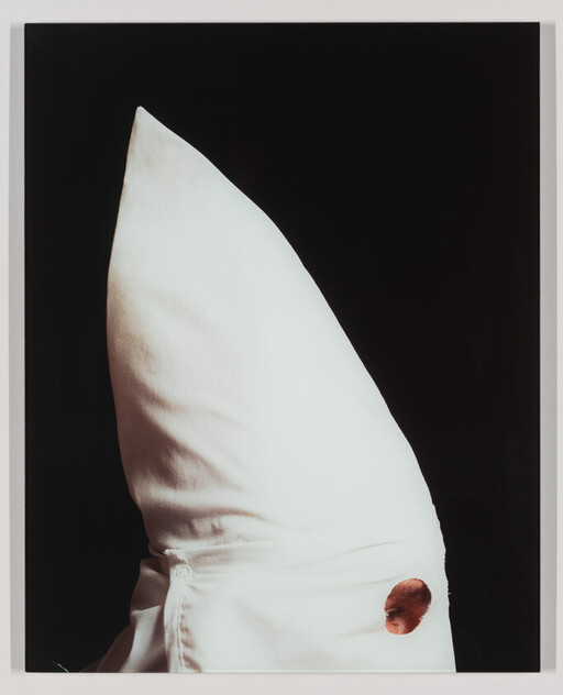 Person wearing a pointed white hood with a single eye hole against a black background.