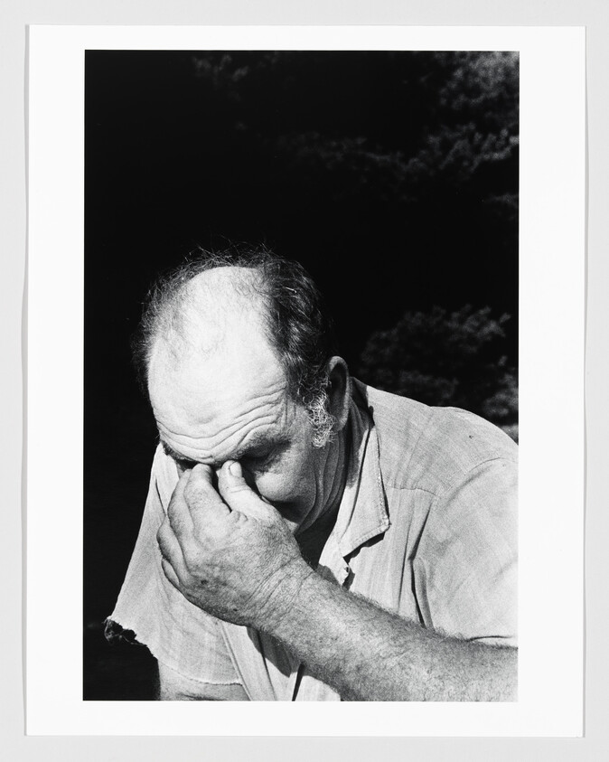 An older balding man leans forward and pinches the bridge of his nose with his hand.