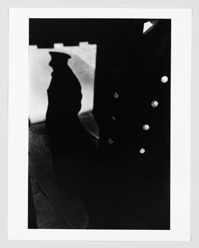 A uniformed figure with shiny buttons casts a strong shadow on a wall.