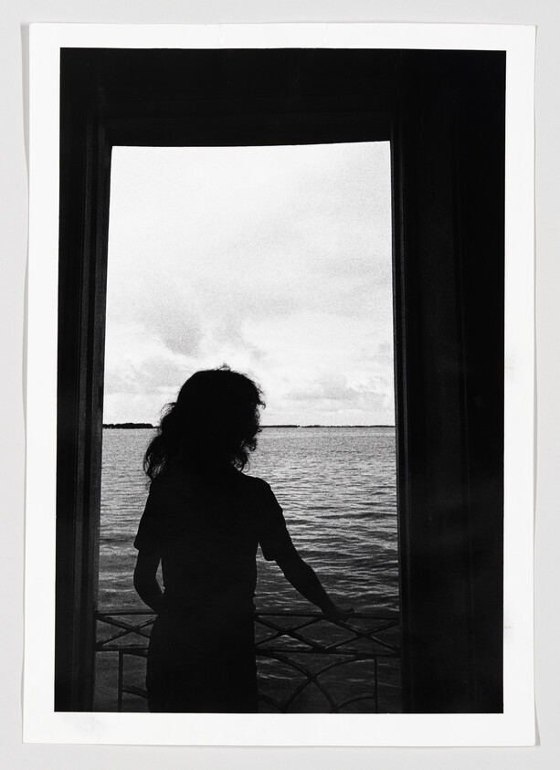 A person silhouetted in a doorway looks out over calm water with hand on railing.