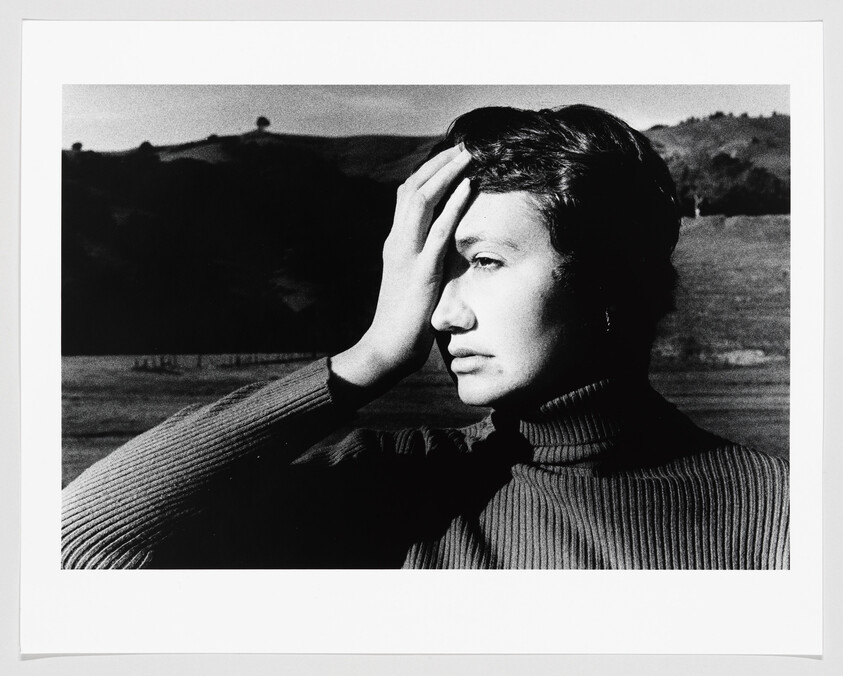 Woman in a turtleneck outdoors holds her hand to her forehead and gazes into the distance.