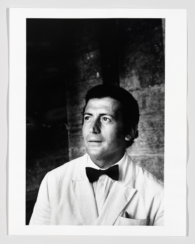 Man in a white suit and black bow tie gazes upward with a thoughtful expression.