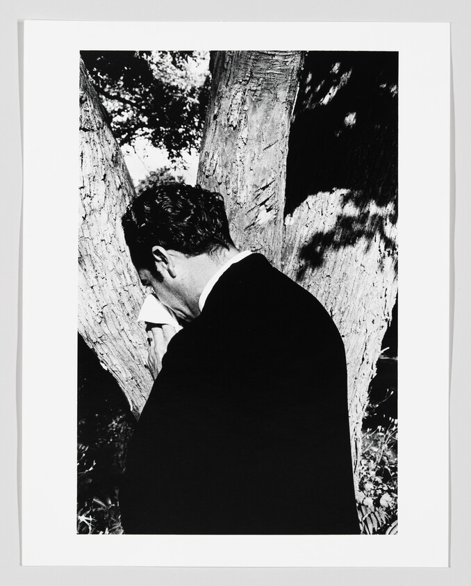 A man in a dark suit stands by a tree wiping his face with a handkerchief.