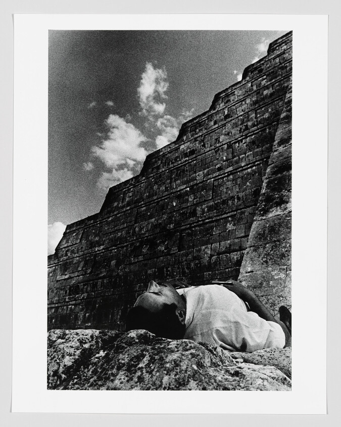 A person lies on rocks at the base of a large stepped stone structure under clouds.