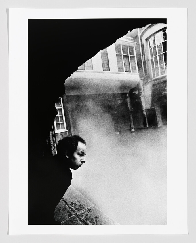 A person leans over a stone ledge peering into thick fog in a courtyard.