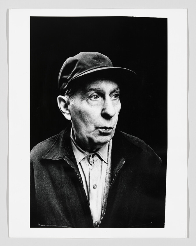 An older man wearing a cap and jacket looks to the side with a surprised expression.