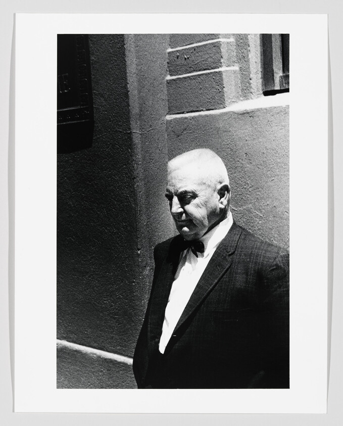 Elderly man in a suit and bow tie stands against a sunlit wall, looking downward.