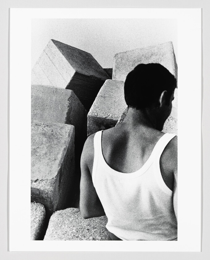 Young man in a white tank top sits with his back to large concrete blocks.