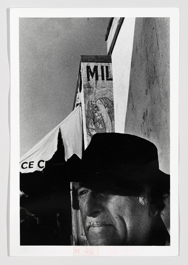 An older man wearing a dark hat stands by a building awning with faded signage.
