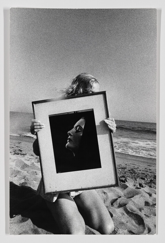 Person kneeling on a beach holds a framed portrait that obscures their face.