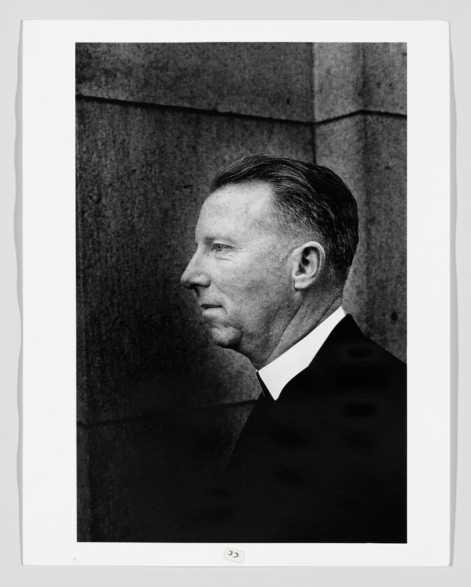 A middle-aged man in a clerical collar shown in left-facing profile against a stone wall.