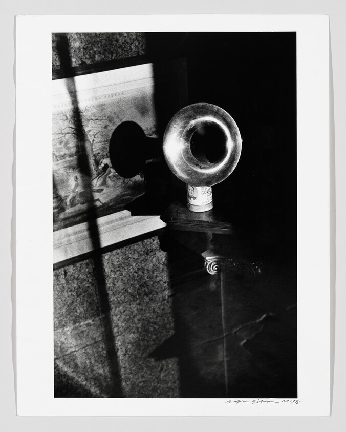 A metal phonograph horn sits on a small pedestal, casting a shadow across a framed picture.