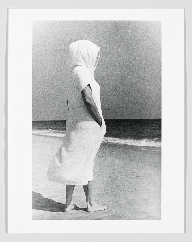 A person in a hooded robe stands barefoot on the beach, hands in pockets, looking at the sea.