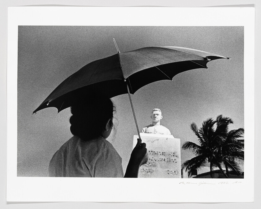 A woman holds an umbrella while looking up at a bust on a pedestal.