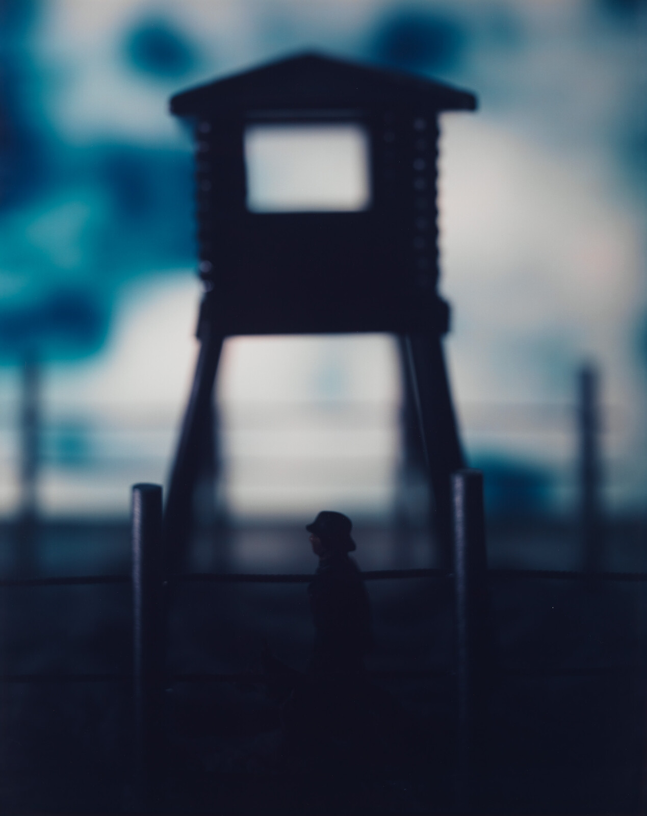Silhouetted guard tower looms over a solitary figure walking by a fence with a dog.