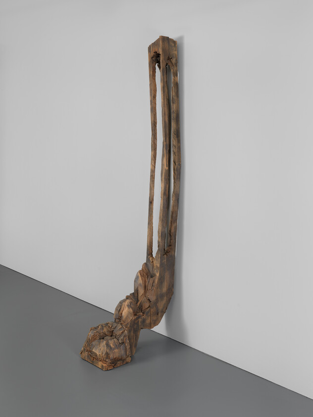 A tall carved wooden sculpture with elongated vertical slats rising from a rough rocky base leaning against a wall.