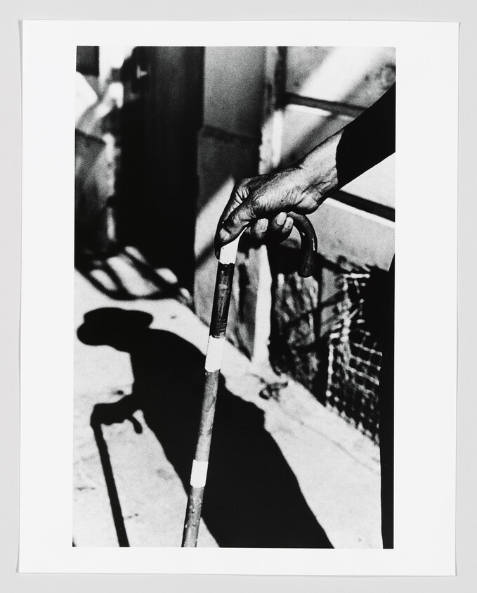 A hand grips a white-tipped cane while its long shadow falls on the sidewalk.