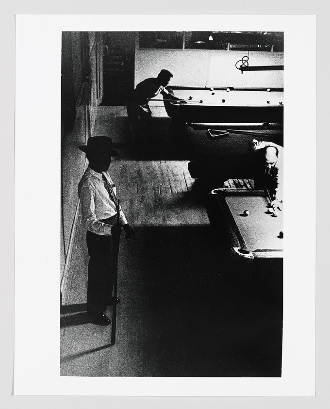 Man in a hat stands with a cue while other players take shots at pool tables.
