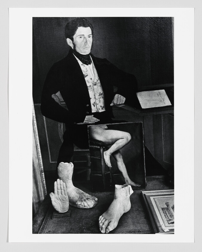 A seated man in formal attire poses behind plaster foot casts and a painting of legs.