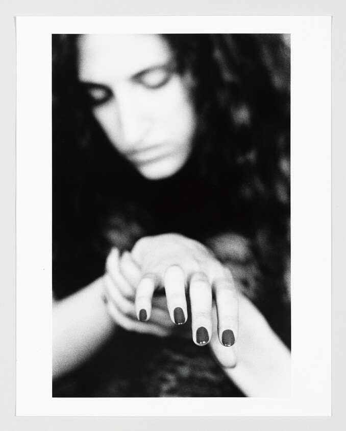 A blurred woman looks down as her hand with dark painted nails reaches forward.