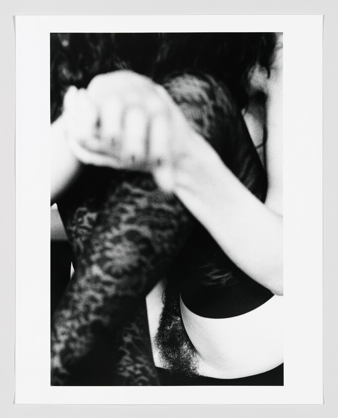 Woman wearing a bra and lace tights sits with arms wrapped around legs, showing natural armpit hair.