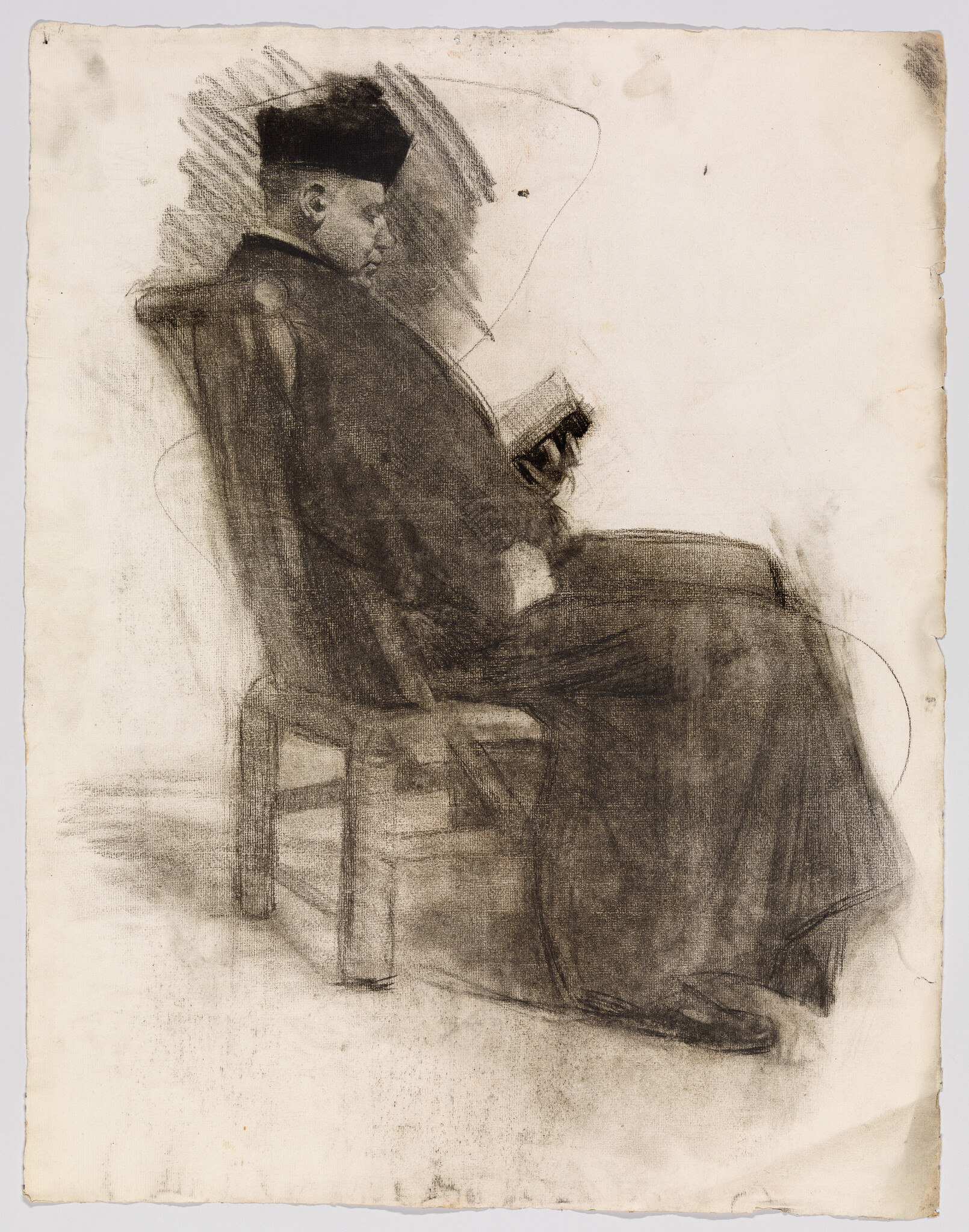 A seated man in a dark robe and hat reads a small book while sitting on a chair.