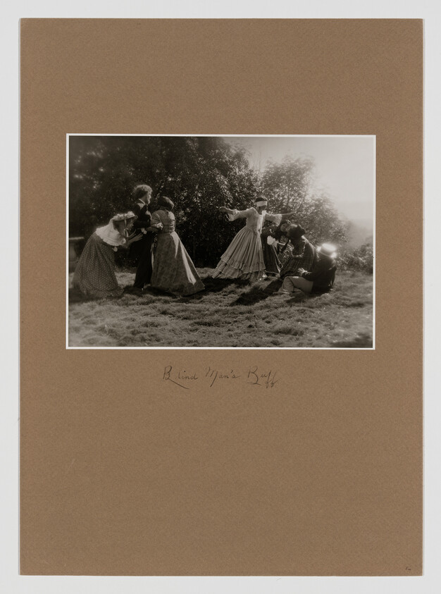 A sepia-toned vintage photograph depicting a group of women in period clothing, possibly from the late 19th or early 20th century, engaging in an outdoor activity. Some women appear to be dancing or in motion, while others are interacting with each other. The setting is a grassy field with trees in the background. The photo is mounted on a brown mat board with a handwritten inscription at the bottom.