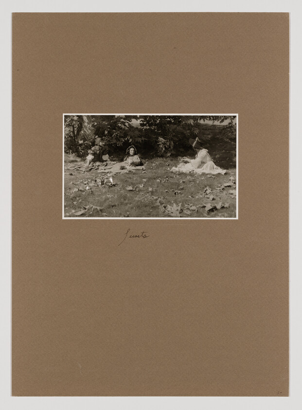 Three women in long dresses sit and relax on a leafy lawn under trees.