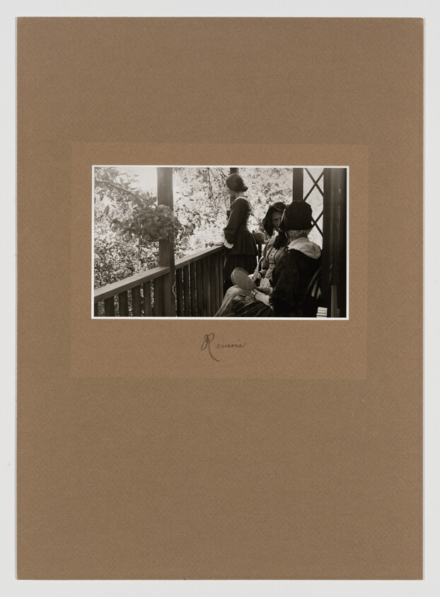Three people in period clothing sit and stand on a porch, surrounded by trees. The scene is captured in a sepia-toned photograph.