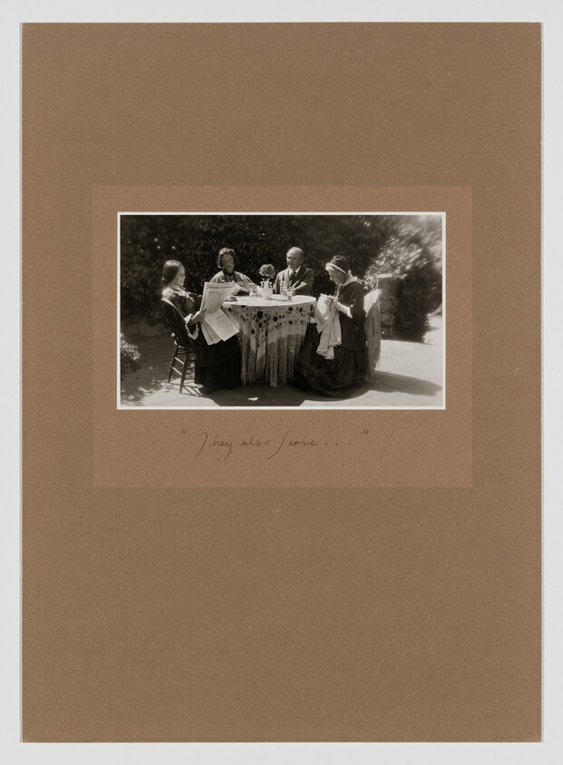 Five people sit around an outdoor table, two women mending cloth while others talk.