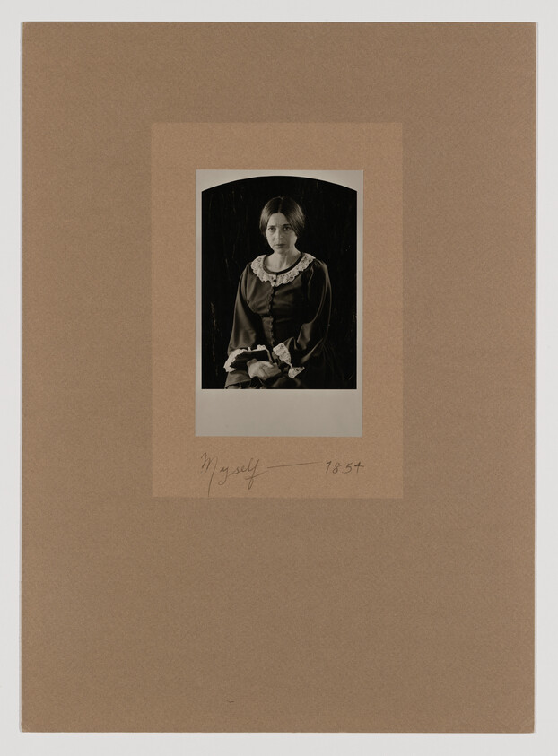 A vintage black and white portrait of a woman mounted on a brown paper mat. The woman is dressed in a dark, old-fashioned dress with a lace collar and is seated with her hands folded in her lap. Below the portrait, handwritten text reads "Myself" followed by the number "7854."