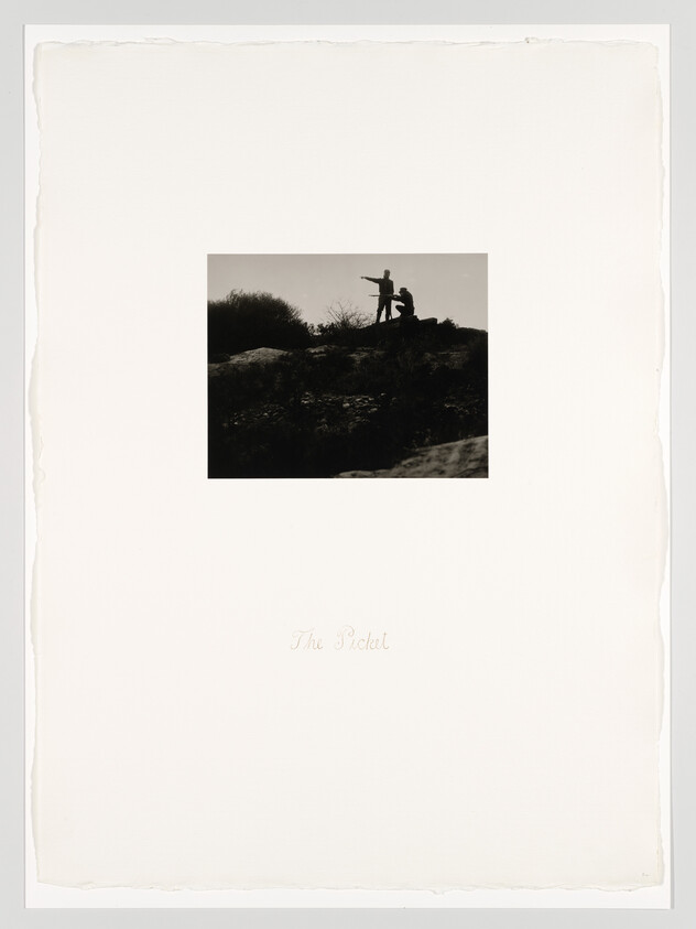 Two people stand on a rocky hill, one pointing while the other kneels beside them.