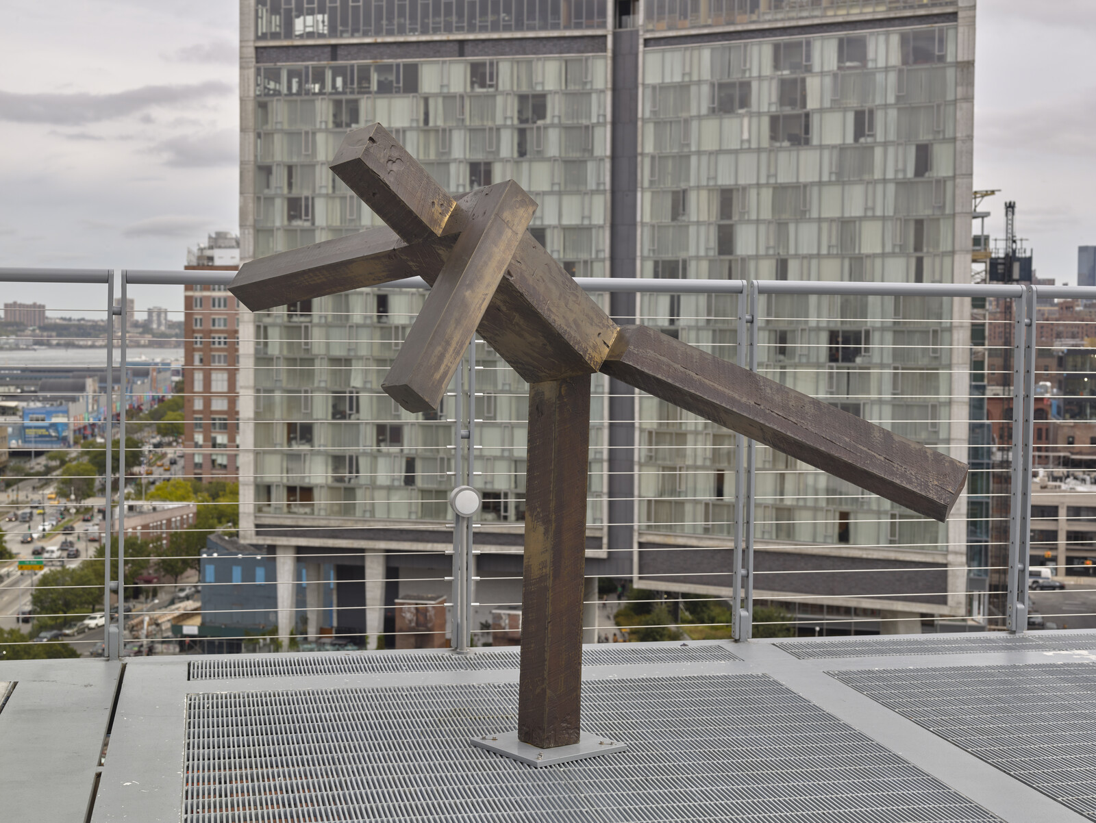 A metal abstract sculpture with intersecting beams stands on a rooftop, with a city building in the background.