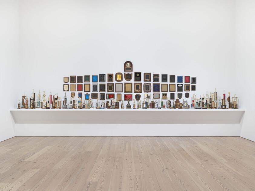 A long shelf holding dozens of trophies and plaques arranged in rows against a white gallery wall.