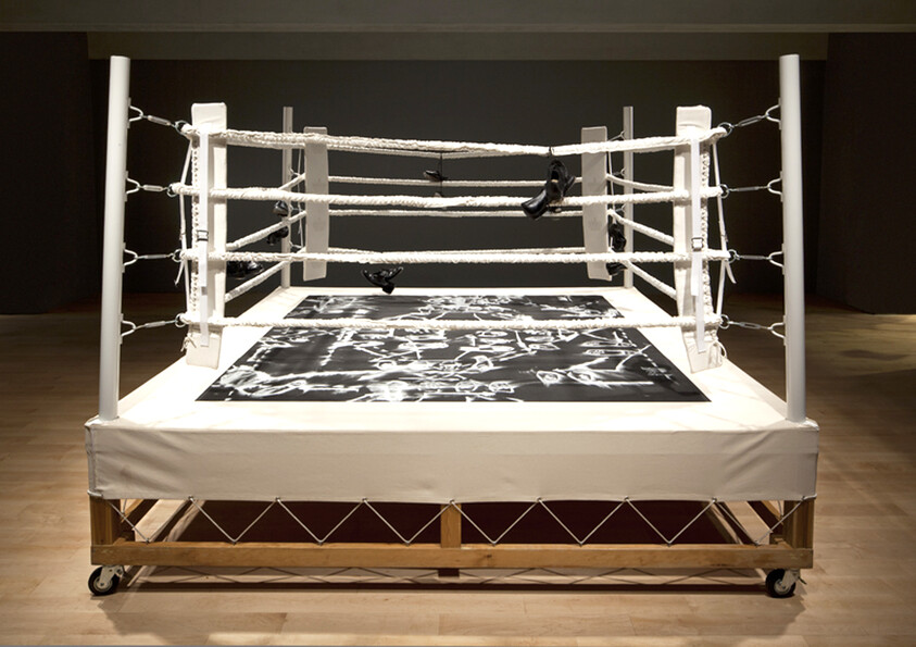 A small white boxing ring on wheels with black-and-white canvas and shoes hanging from the ropes.